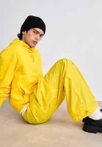 Felpa gialla con cappuccio e pantaloni abbinati in un materiale leggero. Un berretto nero, calzini bianchi texture e scarpe nere chunky completano l'outfit.