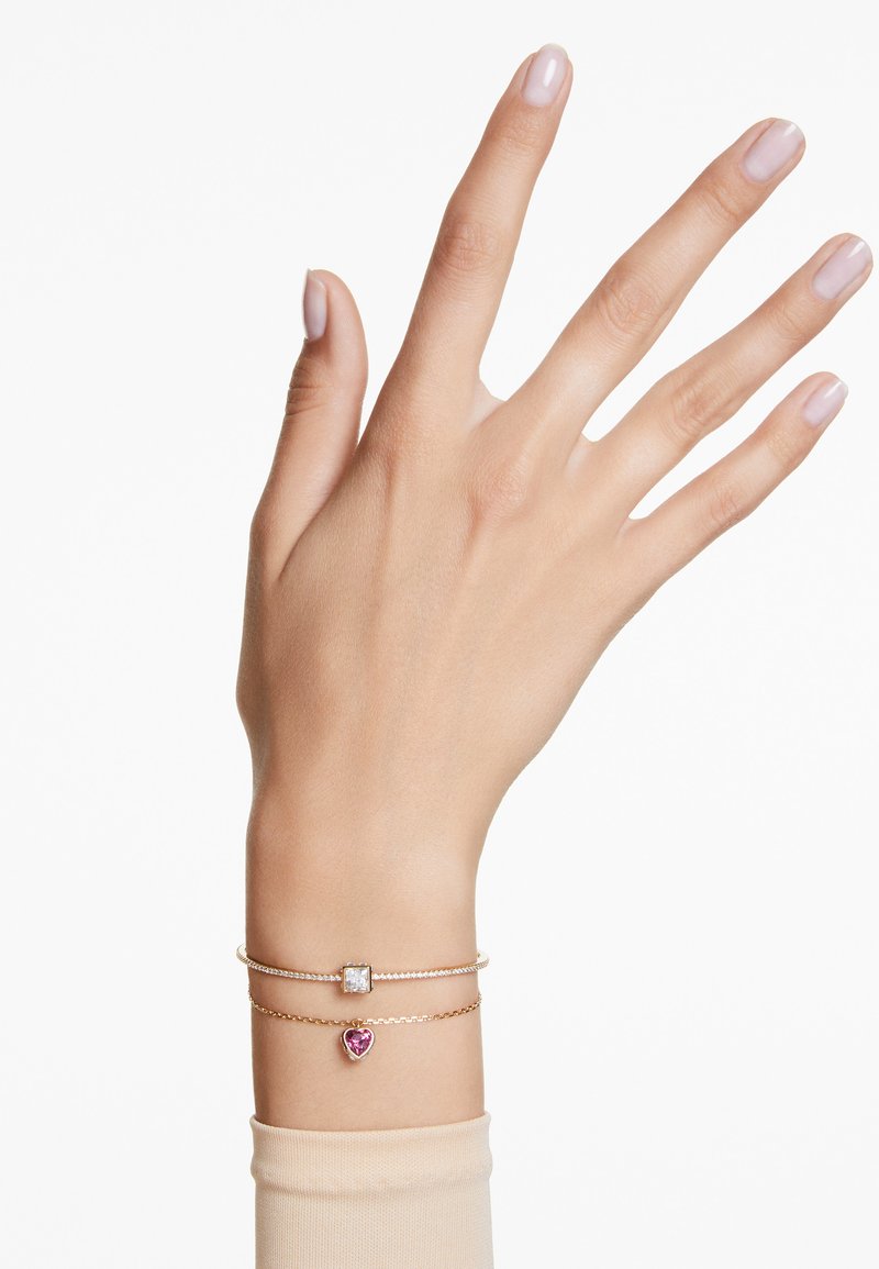 Gold bracelets layering on a wrist, featuring a square clear stone and a pink heart-shaped charm. Smooth texture with slight shine.