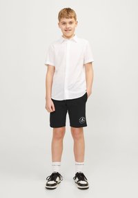 Witte kortere mouw button-up shirt, ontspannen pasvorm zwarte shorts met logo graphic, zwart-witte sneakers en witte crew-sokken.