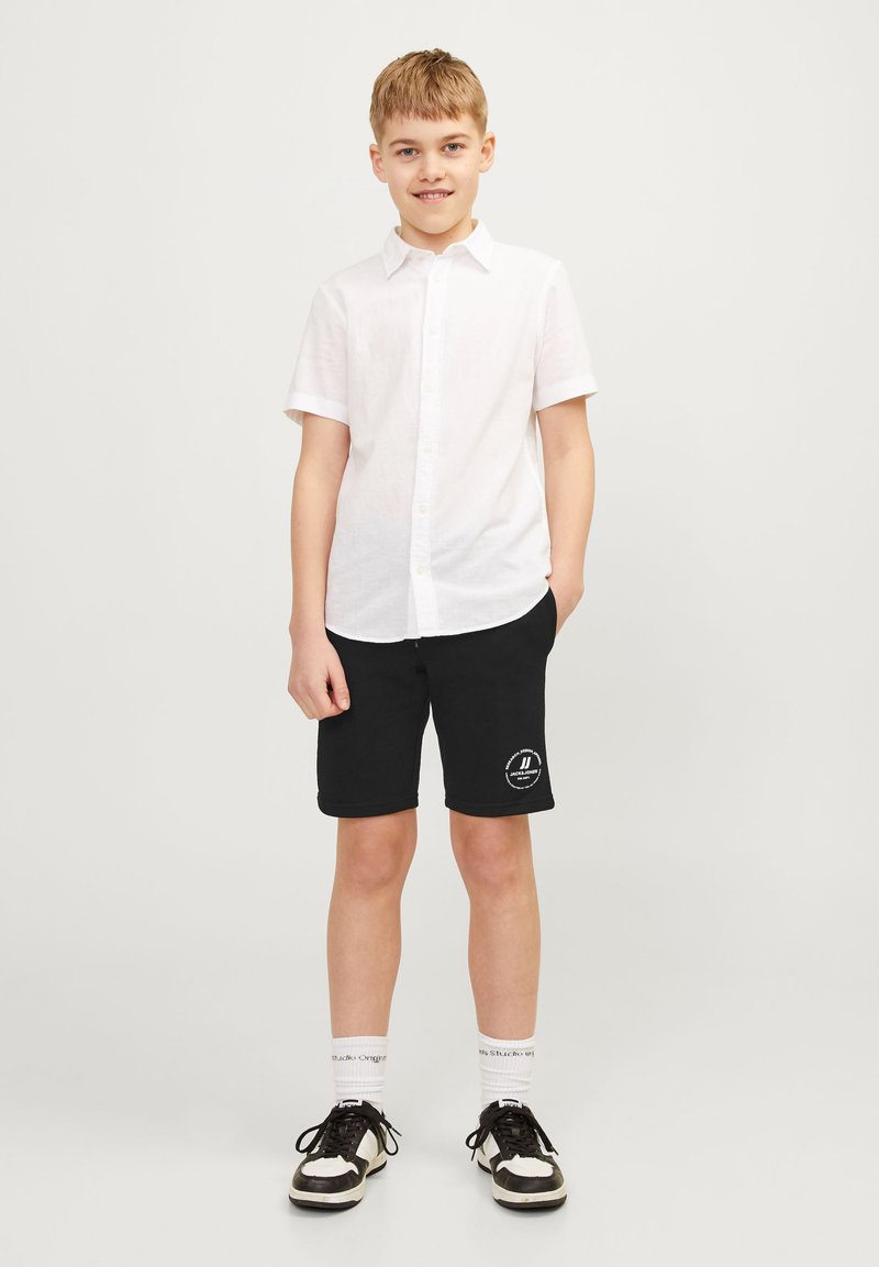 Witte kortere mouw button-up shirt, ontspannen pasvorm zwarte shorts met logo graphic, zwart-witte sneakers en witte crew-sokken.
