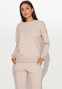 Beżowa bluza z miękkiego materiału, z okrągłym dekoltem, ściągaczami przy rękawach oraz detalem z logo na lewym rękawie. W połączeniu z dopasowanymi spodniami.