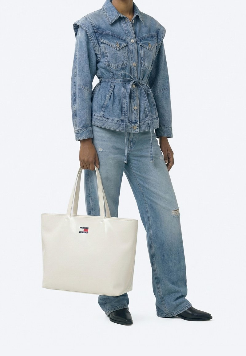 Personne portant une veste en jean bleu clair et un jean, tenant un grand sac cabas blanc avec logo, debout devant un fond uni.
