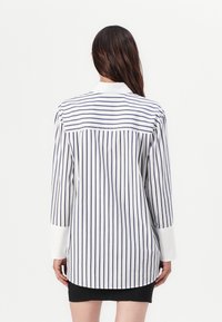 Chemise à manches longues blanche avec des rayures verticales bleu marine, dotée d'un col, de poignets boutonnés et d'un ourlet légèrement arrondi à l'arrière.