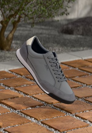 Zapatilla gris de cuero y ante, con diseño texturizado, punta redondeada y cierre frontal con cordones, y suela de goma blanca.