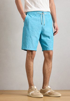 Hellblaue Leinenshorts mit elastischem Bund, Zugband und Seitentaschen. Das Model kombiniert sie mit beigen Sneakers auf einem hellen Holzfußboden.