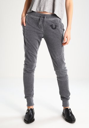 Person trägt dunkelgraue Jogginghosen mit Kordelzug und Logo am linken Oberschenkel, ein kurzes graues Top und schwarze Schuhe mit Reißverschluss, steht mit den Händen in den Taschen.