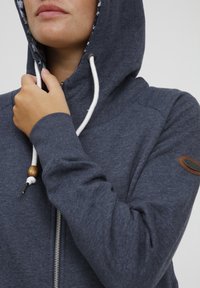 Navy zip-up hoodie with a drawstring hood, featuring a floral inner lining, soft texture, and a brown leather logo patch on the sleeve.