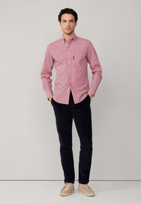 Homme portant une chemise à carreaux rouge à manches longues, un pantalon sombre et des chaussures en daim beige, se tenant devant un fond gris clair uni.