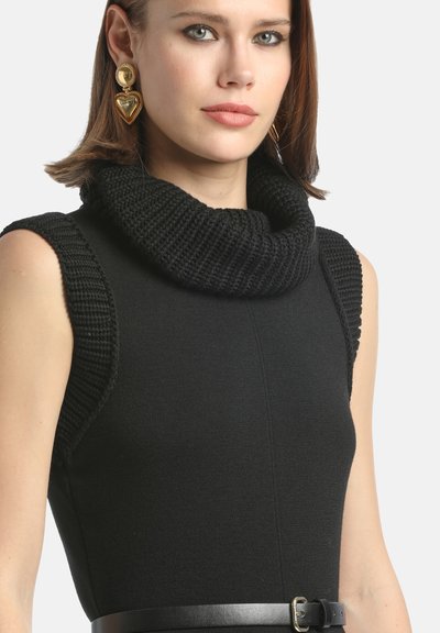 Sieviete, tērpusies bez piedurknēm melnā adītā kleitā ar platu augsto apkakli un melnu jostu, papildināta ar zelta apaļām un sirds formas auskaru. They are a vital accessory.