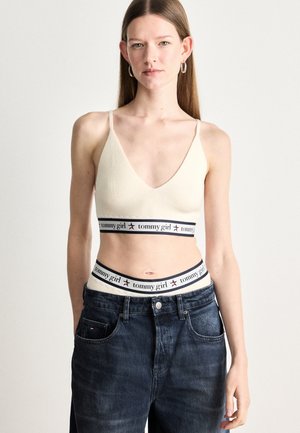 Bralette de canalé crema con tirantes finos, que cuenta con una banda azul marino y blanca etiquetada "tommy girl", combinada con jeans de mezclilla oscuros de tiro alto.