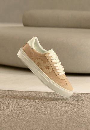 Bézs és fehér sportcipő fehér fűzőkkel és oldalsó logóval, egy texturált szőnyeg felett elhelyezve, háttérben egy kanapéval.