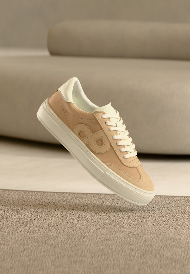 Μπεζ και λευκό αθλητικό παπούτσι με λευκά κορδόνια και λογότυπο στο πλάι, τοποθετημένο πάνω σε υφασμένο χαλί με έναν καναπέ στο υπόβαθρο.