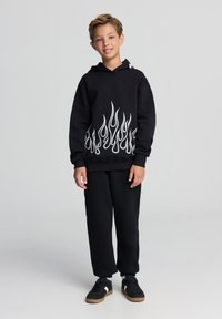 Felpa con cappuccio nera con motivo a fiamma bianca sull'orlo inferiore, abbinata a pantaloni della tuta neri e scarpe da ginnastica nere con suole in gomma.