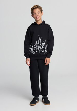 Czarna bluza z kapturem z białym wzorem płomienia u dołu, zestawiona z czarnymi spodniami dresowymi i czarnymi trampkami z gumowymi podeszwami.