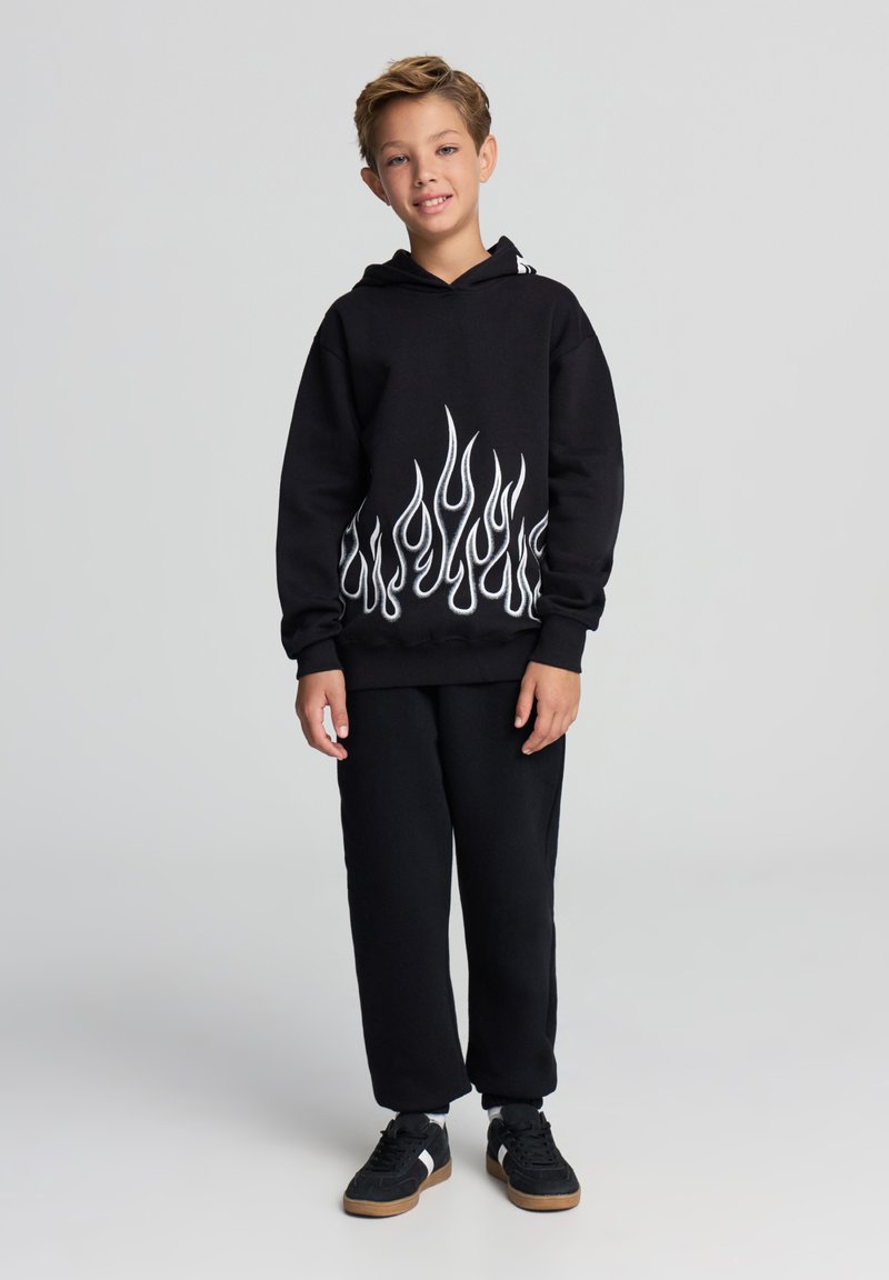 Felpa con cappuccio nera con motivo a fiamma bianca sull'orlo inferiore, abbinata a pantaloni della tuta neri e scarpe da ginnastica nere con suole in gomma.