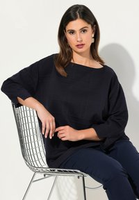 Blusa oversize blu navy con motivo a quadri texturizzati, scollo ampio e maniche a tre quarti, abbinata a pantaloni blu navy. Modella seduta su una sedia in rete.