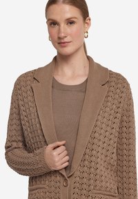 Beige stickad cardigan med ett strukturerat mönster, notchad krage och enstaka knappstängning. Under finns en matchande beige tröja.