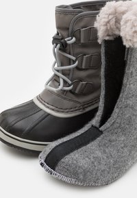 Schwarzer Gummiregenstiefel mit grauem Stoffschaft und Schnürsenkeln, neben einem grauenFilzschläppchen mit schwarzen Stoffakzenten und gesticktem Rand.