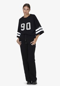Zwart oversized T-shirt met een groot wit detail "90" versierd met studs, gecombineerd met zwarte sweatpants en zwarte schoenen.