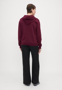 Sudadera burdeos con capucha ajustable, tejido texturizado, corte relajado, combinada con pantalones de pierna ancha negros y calzado gris.