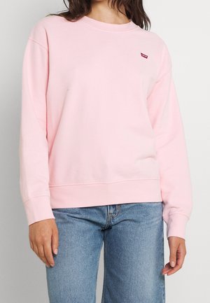 Femme portant un sweat-shirt Levi's rose clair et un jean bleu, debout devant un fond clair uni.