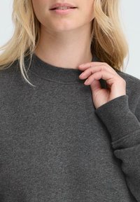 Grå hettegenser med ribbet krage, lange ermer og en myk tekstur. Hånd som hviler på kragen, som viser en avslappet passform.