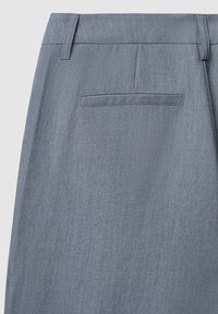 Primo piano di un tessuto grigio chiaro che mostra il retro dei pantaloni con una tasca welt singola e passanti per cintura su uno sfondo uniforme.