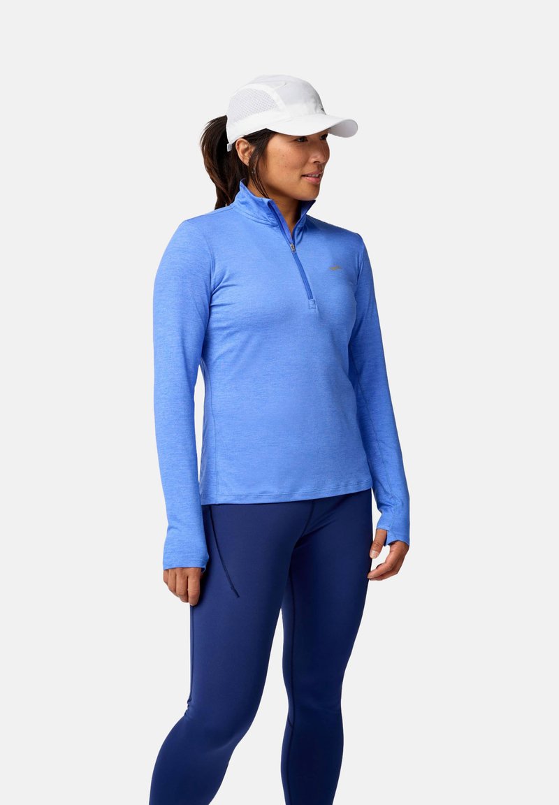 Vaaleansininen pitkähihainen urheilupluuska, jossa on puolivetoedellytys, yhdistettynä tummansinisiin legginsseihin; kangas vaikuttaa kevyeltä ja kosteutta siirtävältä.