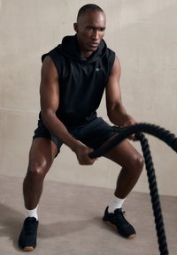 Fekete ujjatlan kapucnis póló, fekete sort és fekete sportcipő, gumitalppal. A személy egy vastag fekete küzdősávot fog meg semleges háttér előtt.