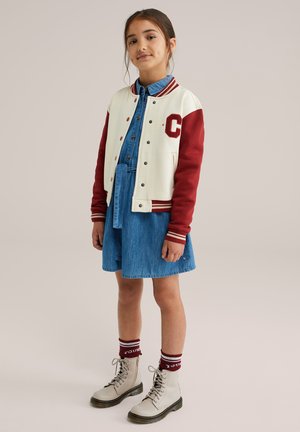 Varsity jaka krēmkrāsā un sarkanā ar 'C' logo; denima pogājamā kleita; krēmkrāsas zābaki ar melniem zole; burgundijas svītrainās zeķes.