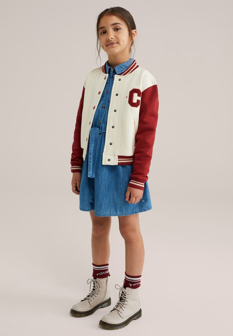 Univerzitetna jakna v kremasti in rdeči barvi z logotipom 'C'; denim srajčna obleka; kremaste čevlje z črnimi podplati; burgundske nogavice z rdečimi črtami.