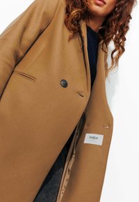 Manteau en mélange de laine brun avec une fermeture à un bouton, deux poches avant et une doublure satinée lisse. L'étiquette indique "GISÈLE."