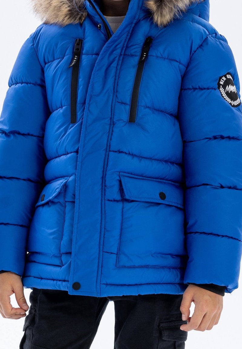 Enfant portant une veste d'hiver matelassée bleue avec capuche bordée de fourrure, fermeture éclair à l'avant, deux poches poitrine et deux poches à rabat, et un pantalon noir.