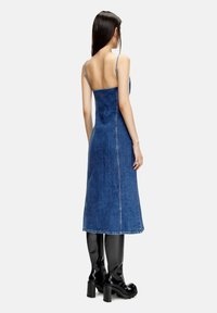 Kvinna klädd i en rygglös, blå denim midi-klänning med tunna axelremmar och svarta högklackade knähöga stövlar, stående framför en neutral bakgrund.