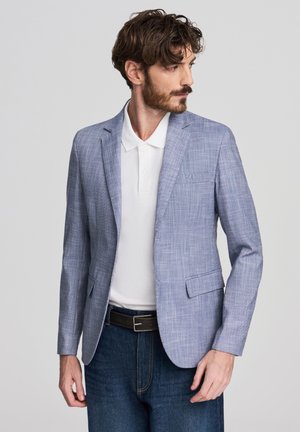Blazer azzurro chiaro con tessuto strutturato, rever a lancia, due tasche frontali e vestibilità sagomata, indossato sopra una polo bianca e jeans scuri.