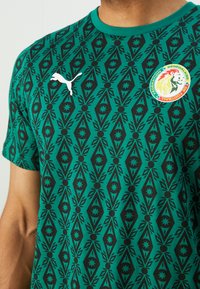 Grünes T-Shirt mit kurzen Ärmeln, das ein schwarzes geometrisches Muster, ein weißes Puma-Logo und ein rundes Senegal-Flaggenabzeichen auf der Brust zeigt.