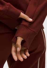 Pullover bordeaux con polsini a foro per il pollice, caratterizzato da cuciture visibili e una superficie liscia. Mani incrociate, una appoggiata sulla manica.