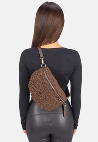 Sac bandoulière en polaire marron avec une surface texturée, une forme asymétrique et un détail de fermeture éclair argenté. Comprend une bandoulière adjustable marron.