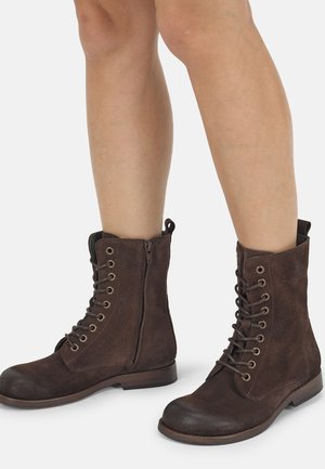 Bottes en daim marron à lacets avec fermeture éclair latérale portées sur jambes nues, montrées de mi-mollet vers le bas sur un fond blanc.