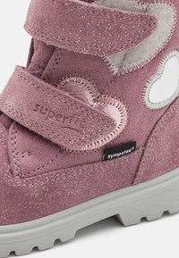 Ροζ σουέτ μπότες με γκλίτερ φινίρισμα, διαθέτοντας δύο λωρίδες Βέλκρο, ζαρωτά στοιχεία σε σχήμα σύννεφου και γκρίζα ελαστική σόλα.  Ορατή ταυτότητα μάρκας.