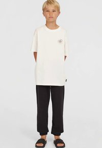 Witte katoenen T-shirt met korte mouwen, voorzien van een cirkelvormige afbeelding op de borst. Gecombineerd met zwarte broek met trekkoord en zwarte sandalen.