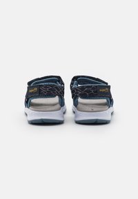 Sandali in tessuto blu con cinturini regolabili, suole bianche e design testurizzato. La vista posteriore mostra un'imbottitura interna morbida e l'etichetta del marchio.