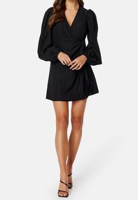 Vestido negro de corte envolvente con rayas verticales texturizadas, escote en V profundo, mangas largas abullonadas y detalle de lazo lateral. Acompañado de tacones negros con tiras.