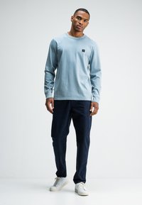 Hellblaues Langarmshirt mit Rundhalsausschnitt und schwarzem Logo-Patch, kombiniert mit marineblauen Cordhosen und weißen Sneakers. Einfaches Design.