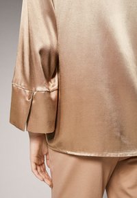 Blusa in satin di bronzo chiaro con manica arrotolata e dettagli con bottoni, texture liscia, con una vestibilità leggermente morbida. L'orlo cade sotto la vita.