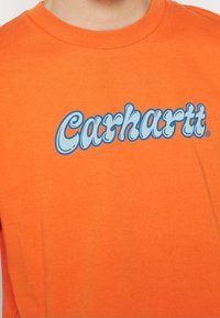 Bright orange t-shirt with light blue and dark blue outlined "Carhartt" text logo on the chest, worn by an upper torso and neck.