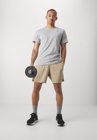 Camiseta atlética gris, pantalones cortos beige, sosteniendo un disco de pesas negro. Zapatillas atléticas negras con calcetines grises. Diseño simple y funcional.
