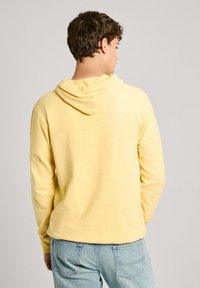 Sudadera amarilla de corte holgado, con mangas largas y capucha. Fabricada con una tela suave de textura sutil. Usada con jeans de color azul claro.