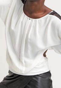 Witte blouse met plooien en kantdetails op de schouders, met een relaxte pasvorm en een zachte textuur. Gecombineerd met een zwarte leren onderkant.
