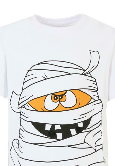 T-shirt en coton blanc avec un dessin de momie en cartoon, au visage orange, avec de grands yeux et un bandage rayé noir et blanc, ainsi que des accents de boutons.